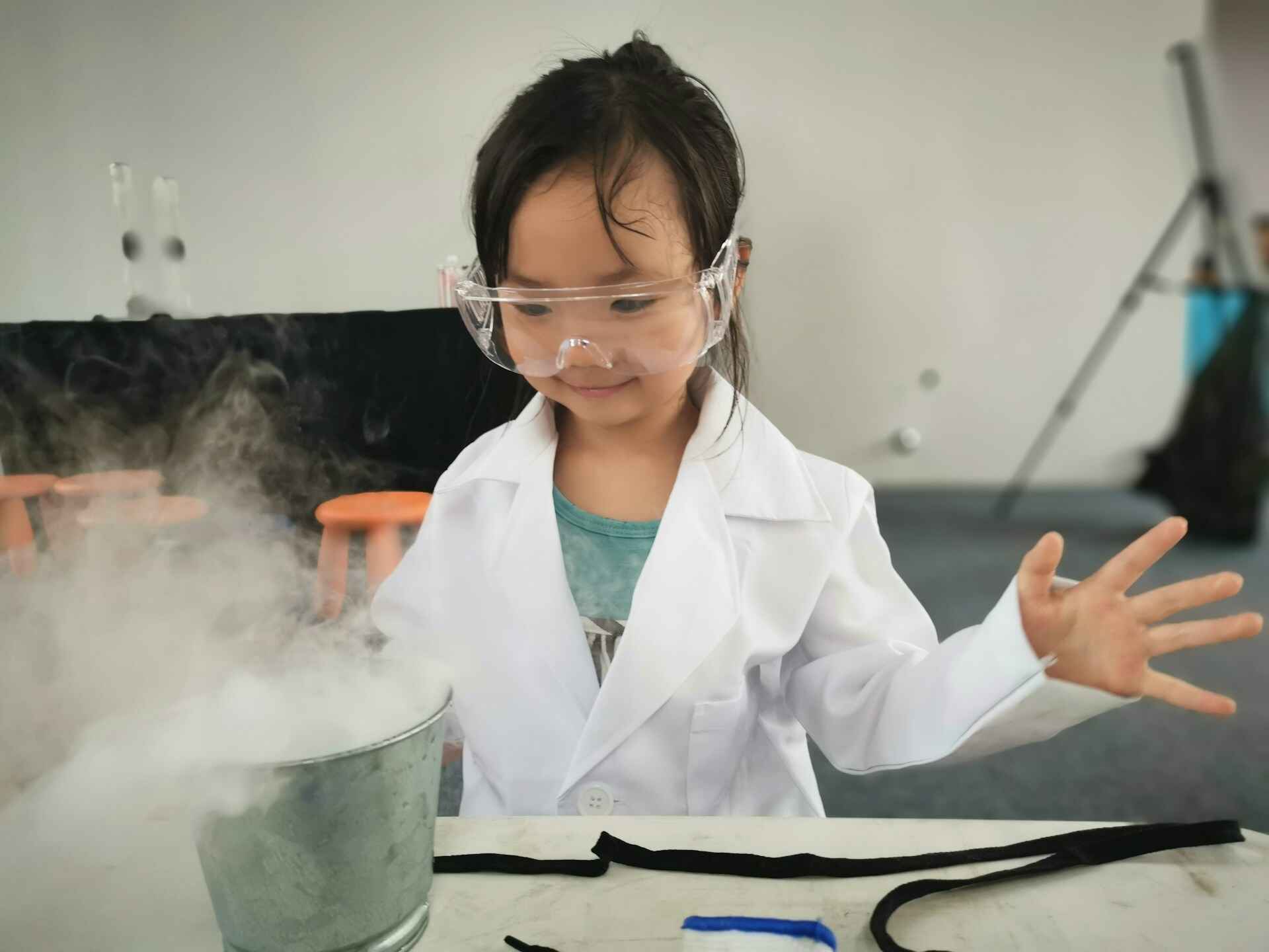 A child in a lab coat and protective clear glasses looking at a bucket of liquid nitrogen smoke pourig out of it